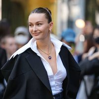 Guest wears a black structured blazer draped over the shoulders, paired with a crisp white button-up shirt featuring a wide collar. The hair is...