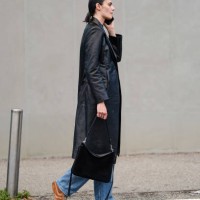 Guest wears a black t-shirt, a black leather trench coat, a black leather bag, blue denim jeans / pants, brown suede sneakers shoes, outside Jil...