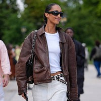 Guest wears a brown leather jacket with a classic collar and zip fastening. Underneath, a white fitted t-shirt is visible. The outfit includes white...