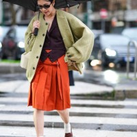 Guest wears a burgundy waistcoat, a green checked oversized jacket, sunglasses, a white leather bag, an orange midi pleated skirt, brown leather...