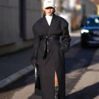Guest wears a gray cap hat, a white turtleneck pullover , a black long winter coat, a belt, knee high black leather boots , a leather bag, outside...