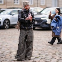 Guest wears a gray fluffy chapka hat, a black puffer jacket, flared pants , beige shoes, holds a mobile phone to take a picture / selfie, outside...