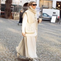 Guest wears a long white pleated maxi skirt, white jacket, white knitted vest, beige bag, and grey sneakers outside Herskind during the Copenhagen...