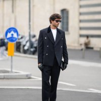 Guest wears a military / blazer jacket, a white t-shirt, black leather gloves, black pants, black shiny leather pointed shoes, outside Prada, during...