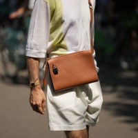 Guest wears a pale gray with large pale khaki strap pattern t-shirt, a camel shiny leather crossbody bag, beige suit shorts, a gold watch , outside...