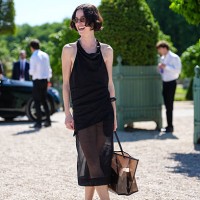 Guest wears a sleeveless black dress with a draped neckline and a sheer overlay extending below the knee. The hair is styled in a short, wavy bob....