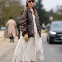 Guest wears a white gathered dress with low-neck and floral embroidery, a brown oversized leather jacket, beige gloves, sunglasses, black ballet...