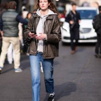 Guest wears a white / grey hoodie, a brown leather jacket, blue denim jeans / pants, a black leather bag, black and white sneakers, outside Onitsuka...
