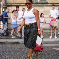 Guest wears a white ribbed tank top paired with dark denim knee-length shorts featuring prominent stitching and metal embellishments. Accessories...