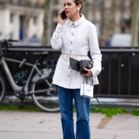 Guest wears a white tweed coat, a flower shaped earring, a black Chanel bag, blue denim jeans / pants, white ballerina shoes outside Chanel, during...