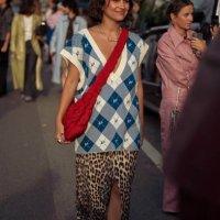 Guest wears animal print long skirt with split, white and blue BP checkered vest and red shoulder bag outside the Baum und Pferdgarten show during...