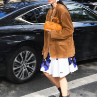 Guest wears beige Miu Miu top, white Miu Miu floral print skirt, brown suade blazer, orange Miu Miu bag, grey socks, brown loafers, outside Miu Miu...