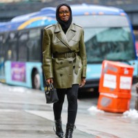 Guest wears black balaclava, black sunglasses with orange lenses, shiny olive green belted double breasted leather coat from Prada, black mini...