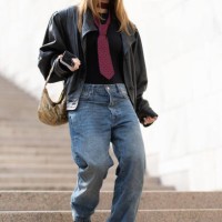 Guest wears black leather heeled boots, a yellow leather bag, brown sunglasses, a dark red printed tie, a black t-shirt, a dark brown leather bag and...