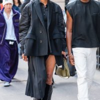 Guest wears black shirt, black jacket with blazer details, black pleated skirt, black boots, outside Sacai, during the Haute Menswear Spring/Summer...