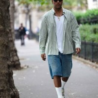 Guest wears black small sunglasses, earrings, a white t-shirt, a white and green striped print pattern shirt, gold chain pendant necklaces, blue...