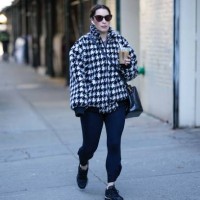 Guest wears black sunglasses, a black and white houndstooth print pattern oversized bomber coat, a black shiny leather crossbody bag, black legging...