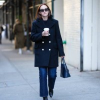 Guest wears black sunglasses, a black t-shirt, a black buttoned coat, a navy blue shiny leather handbag, navy blue pants, black shiny leather pointed...