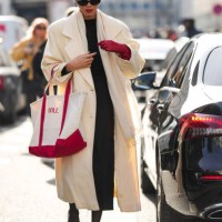Guest wears black sunglasses, a black wool pullover, a red shiny leather high glove, black short legging pants, a white latte oversized long coat, a...