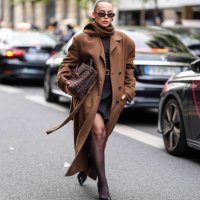Guest wears black sunglasses, a brown scarf, a brown oversized coat, a dark brown top, a black mini skirt, black tights, brown leather bag, black...
