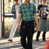 Guest wears black sunglasses, a green with embroidered black pattern short sleeves polo shirt, a black shiny leather belt, black large suit pants,...
