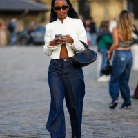Guest wears black sunglasses, a white high neck / jacket, a black shiny leather handbag, navy blue denim wide legs pants, black velvet ballerinas ,...