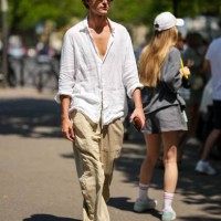 Guest wears black sunglasses, a white linen cotton shirt, beige large pants, dark brown shiny leather shoes , outside Hermes, during the Menswear...