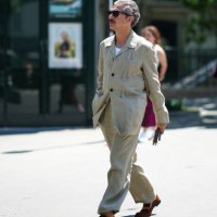 Guest wears black sunglasses, a white t-shirt, a beige buttoned with cut-out leaves pattern jacket, matching beige linen large pants, orange camel...