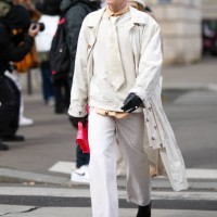 Guest wears black sunglasses, a white tie, a white latte sweater, a white long belted coat, white denim large pants, black shiny leather gloves, a...