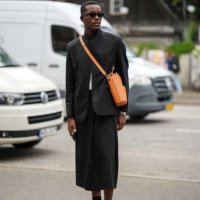 Guest wears black sunglasses, an orange shiny leather crossbody bag, a black blazer jacket, a white t-shirt, a black midi skirt, black socks, yellow...