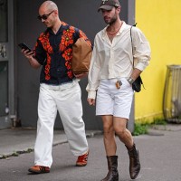 Guest wears black sunglasses, black bright cherry red floral pattern/print buttoned up short sleeve shirt, dark brown leather toe bag, white baggy...