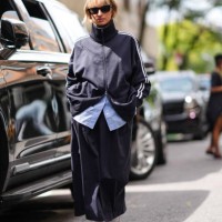 Guest wears black sunglasses, black high track suit jacket, light blue buttoned up long sleeve shirt, black midi skirt, white socks, shiny red...