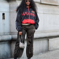 Guest wears black sunglasses, black red graphic oversized jacket, dark green baggy pants, light gray pattern/print leather bag, white socks, red...