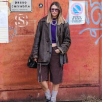 Guest wears black sunglasses, dark purple sweater, white layered pearl necklaces, dark gray oversized blazer jacket, shiny dark brown leather jacket,...