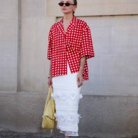 Guest wears black sunglasses, gold earrings, white and red pattern button up short sleeve shirt, silver bracelet, light yellow leather bag, white...