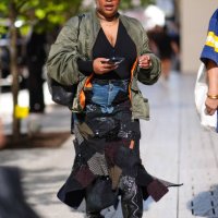 Guest wears black sunglasses, gold necklace, black v-neck shirt, dark green bomber jacket, navy blue denim jean mixed patch midi skirt, shiny black...