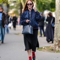 Guest wears black sunglasses, navy blue bomber jacket, shiny dark navy blue leather bag, black satin midi skirt, bright red socks, shiny black...