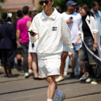 Guest wears black sunglasses, necklaces, a white latte with embroidered black logo pattern long sleeves / polo neck sweater from Miu Miu, white sport...