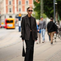 Guest wears black sunglasses, silver earrings, a black cropped top, a black oversized blazer jacket, a black high waist long skirt, a black shiny...
