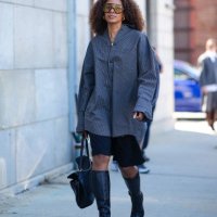 Guest wears black sunglasses, silver earrings, dark gray oversized long sleeve shirt, silver rings, shiny black leather bag, black shorts, shiny...