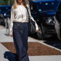 Guest wears black sunglasses, silver necklace with pendant, cream lustrous silky long sleeve shirt, dark navy blue denim loose pants, bright red...