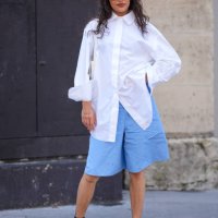 Guest wears black sunglasses, white oversized button up long sleeve shirt, white leather bag, light blue shorts, black socks, shiny black loafer...