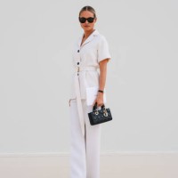 Guest wears black sunglasses, white short sleeve belted buttoned up blazer jacket, white straight trouser pants, shiny black quilted Dior Lady D-Joy...