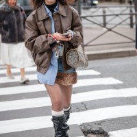 Guest wears blue shirt, brown leather jacket, denim mini skirt with fur detail, black boots, brown bag, outside Miu Miu, during the Womenswear...