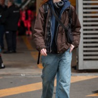 Guest wears blue sweatshirt, brown jacket, jeans, black crossbody bag, outside Ferragamo, during the Milan Fashion Week - Womenswear Fall/Winter...