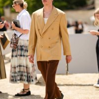 Guest wears brown and orange sunglasses, a white t-shirt, a yellow buttoned long blazer jacket, dark brown velvet suit pants, black shiny leather...