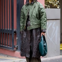 Guest wears brown leather trench coat, olive green pants, brown boots, olive green jacket, green Bottega Veneta bag, black Sunnei sunglasses, outside...