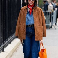 Guest wears brown suede jacket, navy blue denim jeans, orange bag, blouse, red scarf outside Lacoste during the Womenswear Fall/Winter 2025/2026 as...