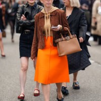 Guest wears brown sunglasses, a brown suede jacket, a light brown hoodie, a brown Prada leather handbag, a long flashy orange skirt, black pointed...