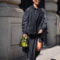 Guest wears brown sunglasses, silver earrings, black button up long sleeve shirt, black shiny black blazer bomber jacket, a light green leather bag...